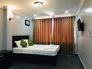 A hotel room with a bed, a nightstand, a TV mounted on the wall, and a window with curtains. - Maison Rivera Guest House (Kampot)