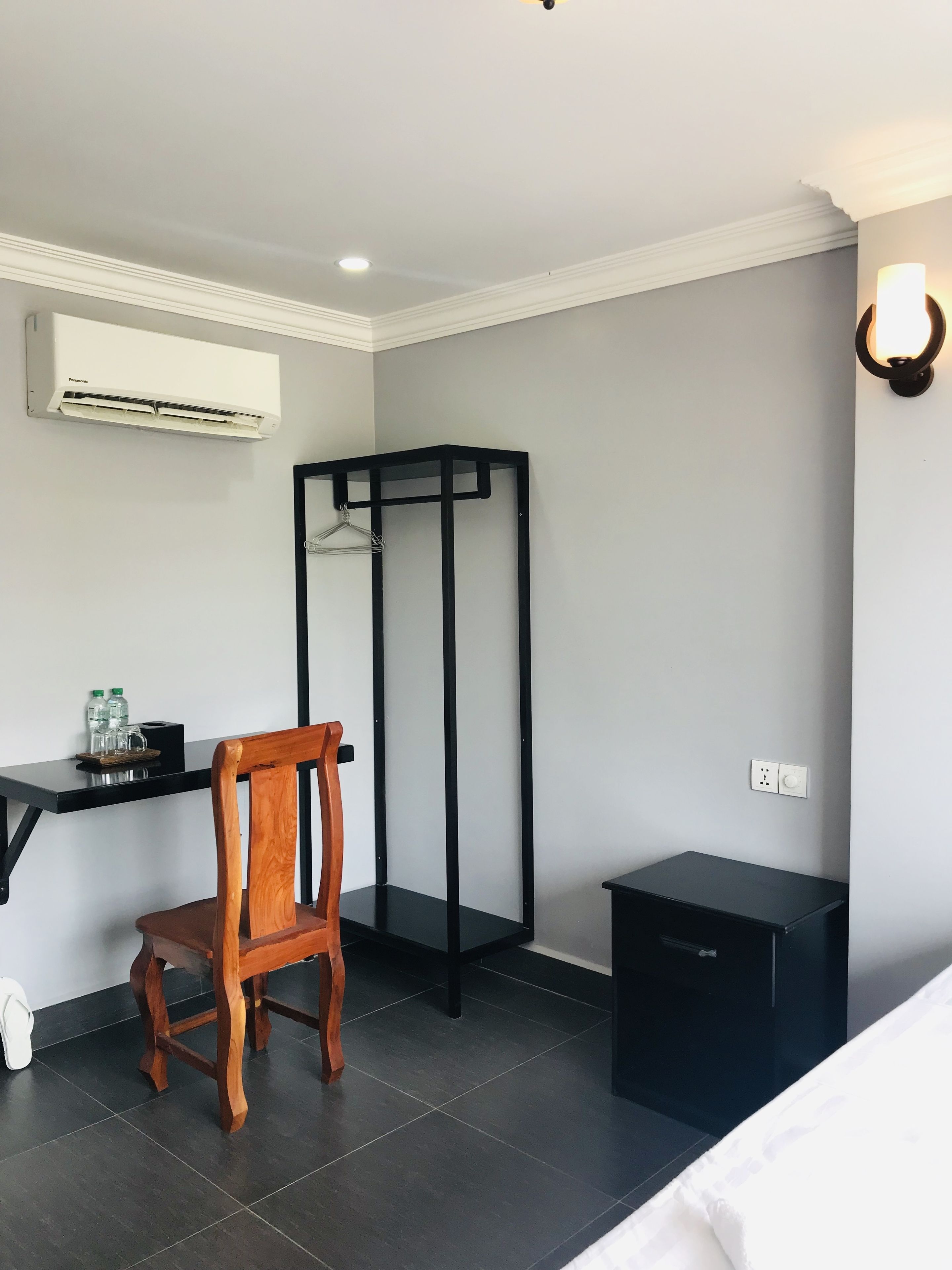 A hotel room with a wooden chair, a desk with a black metal frame, a black storage unit, and a white air conditioning unit on the wall.