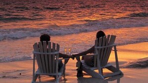 Two people sitting on Adirondack chairs facing the ocean at sunset.