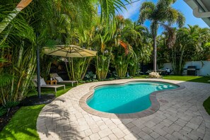 A swimming pool with a curved edge, surrounded by a paved patio and lush tropical vegetation.