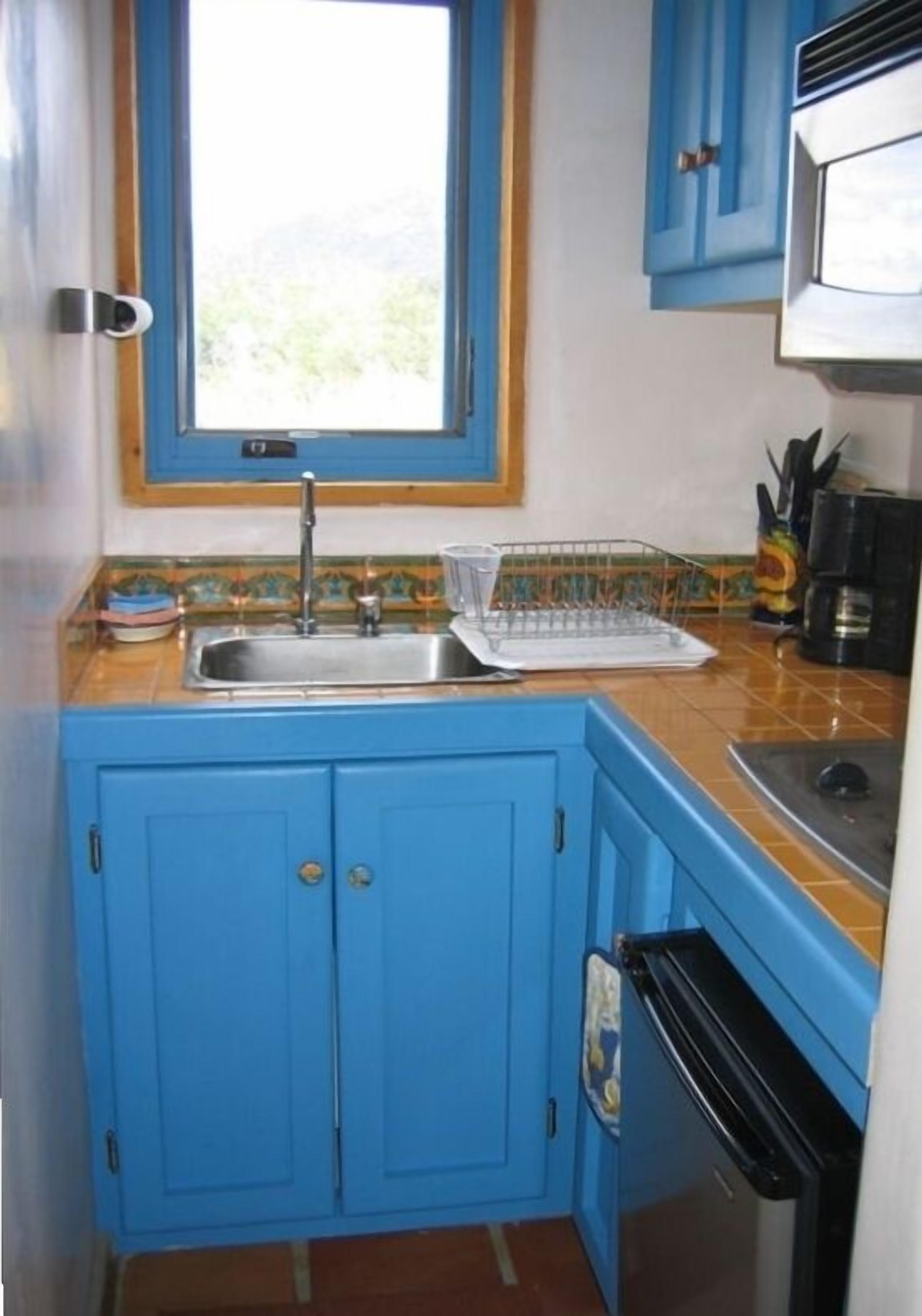 Cocina pequeña con armarios azules, un lavamanos de acero inoxidable y una ventana sobre el lavamanos.