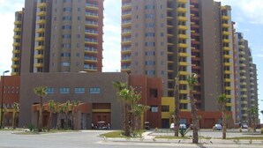 Un immeuble résidentiel à plusieurs étages, doté de balcons, d’un espace de stationnement et de palmiers.