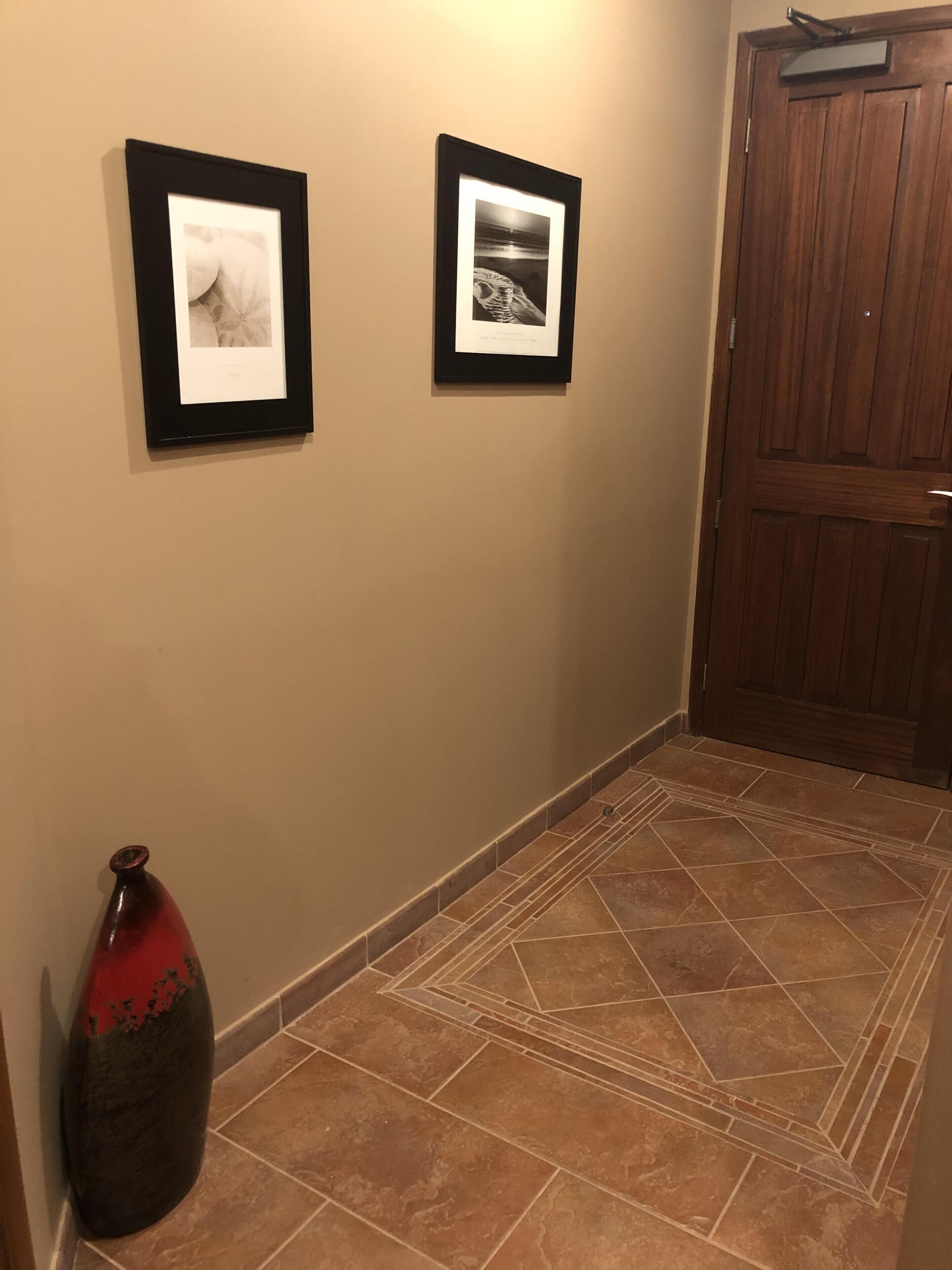 A hallway with two framed photographs on the wall, a wooden door, and a tiled floor.