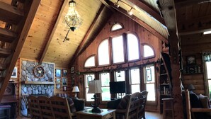 A cozy cabin interior with wooden walls, a stone fireplace, a wooden dining table, and a large window. - Beautiful Beachfront Custom Log Cabin on Lake Huron (Oscoda)