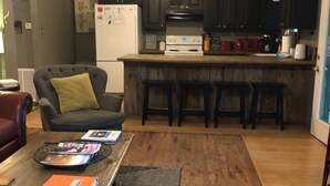 Cocina con armarios oscuros, un refrigerador blanco, un microondas y una estufa. Hay una zona con taburetes de bar y una mesa de comedor con revistas y un cuaderno.