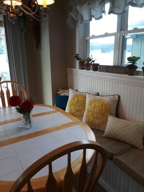A cozy dining area with a round table, chairs, a vase with flowers, and a window with potted plants.