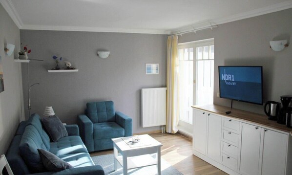 A modern living room with a sofa, armchair, and coffee table. There is a TV on the wall and a radiator. - (17) 2-room apartment Ostseeallee - Appartmenthaus Sonnenburg (Kühlungsborn)