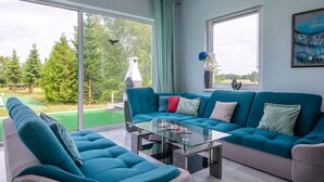 Un salon moderne avec un canapé turquoise, une table basse en verre et une vue sur un jardin et des arbres, grâce à de grandes fenêtres.