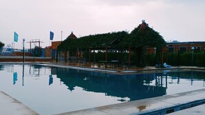 Una piscina con pabellón y zona cubierta, rodeada de vegetación y banderas.