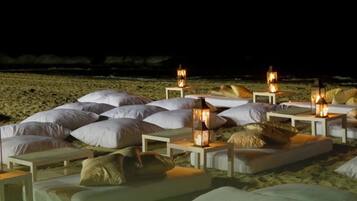 A beachside lounge area with multiple white cushioned seating areas, each with a small table and a lantern.