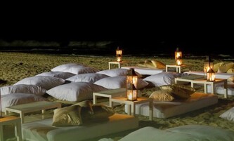 Un espace de détente aménagé au bord de la plage, avec plusieurs coins salons dotés de sièges blancs et moelleux, chacun comprenant une petite table et une lanterne.