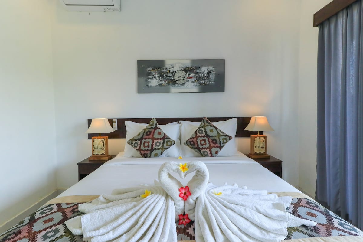 Een netjes opgemaakt bed met handdoeken in de vorm van een hart, twee decoratieve kussens, nachtlampjes en een aan de muur bevestigd kunstwerk.