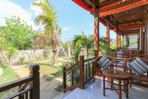 A covered outdoor seating area with wooden furniture and a view of a garden with palm trees. - Kayu Manis Guest House Lembongan (Lembongan Island)