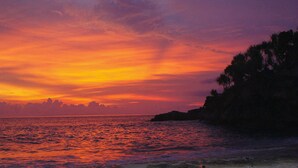 Un coucher de soleil sur une zone côtière, avec une vue sur la mer et la silhouette d’arbres sur la droite.
