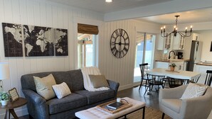 A cozy living room with a gray sofa, a coffee table, and a dining area with a white table and chairs. A large wall clock and world map artwork are visible. - Updated charming home - perfectly located between Downtown & Kidsport Complex (Kalispell)