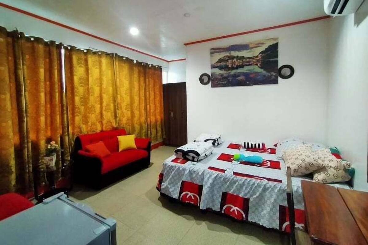 A room with a red sofa, a bed with a red and white bedspread, a small table, and a wall-mounted air conditioner.