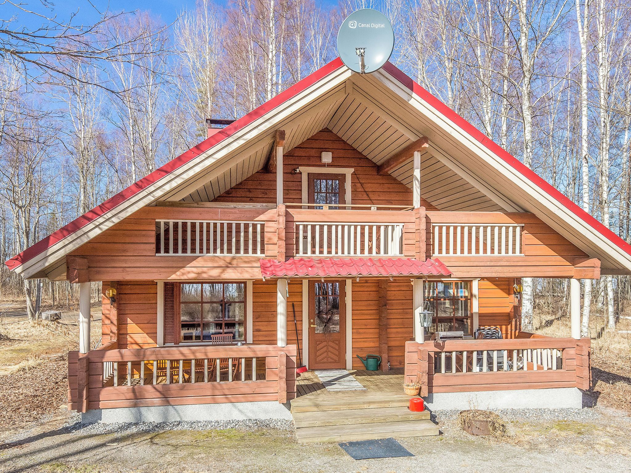 A two-story log cabin with a red roof, a balcony on the upper floor, and a satellite dish on the roof.