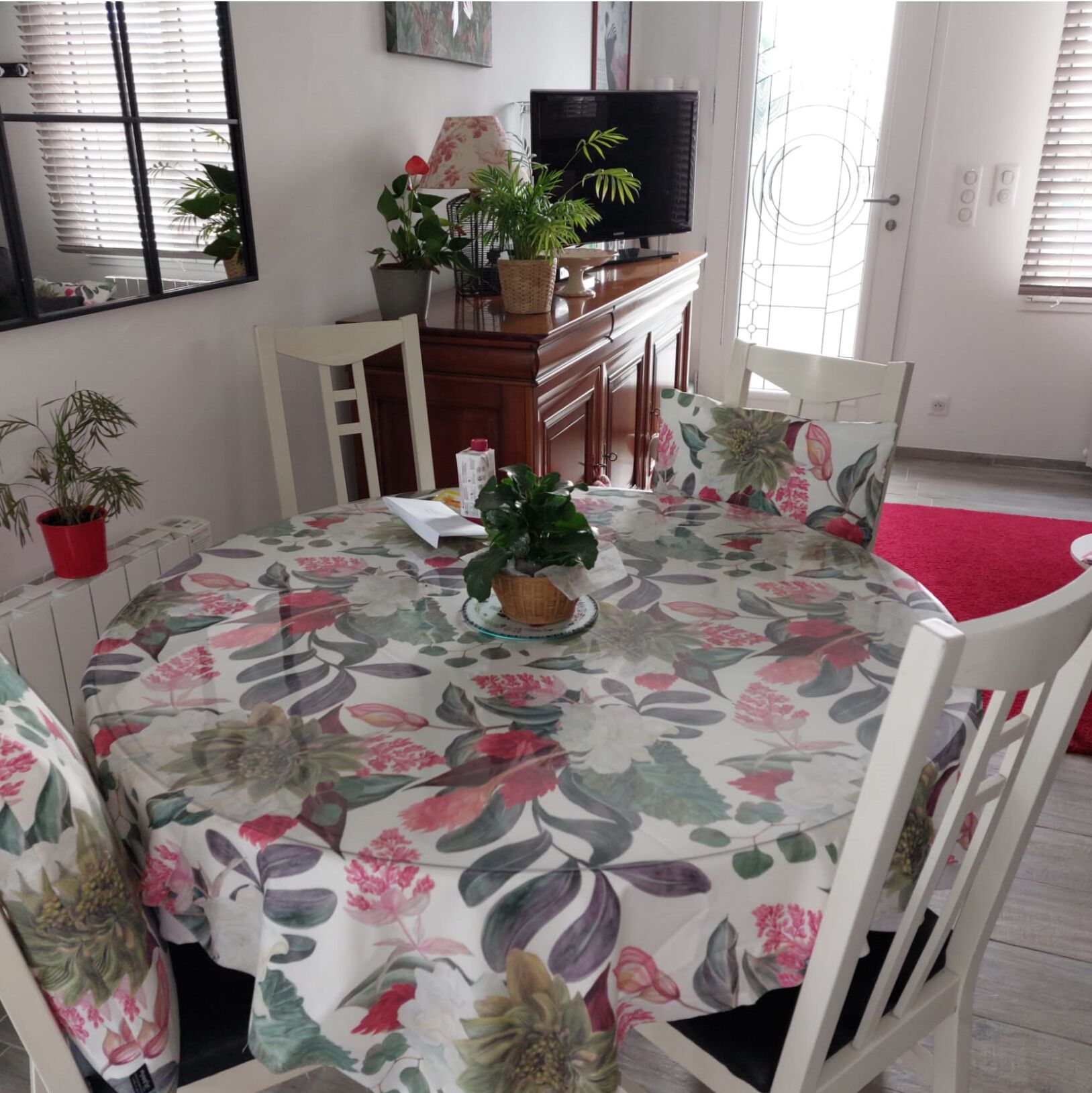 Un coin repas avec une nappe à motifs floraux, des chaises blanches, un buffet en bois et un téléviseur posé sur un meuble.