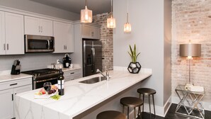 Une cuisine moderne avec des meubles blancs, un réfrigérateur en acier inoxydable, un four à micro-ondes encastré et un comptoir en marbre. On y trouve des luminaires suspendus, une plante en pot et un coin bar avec des tabourets.