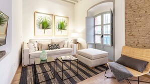 Una sala de estar con sofá, reposapiés, mesa de centro y una silla de mimbre. Hay dos cuadros enmarcados, una lámpara y una ventana con vistas.