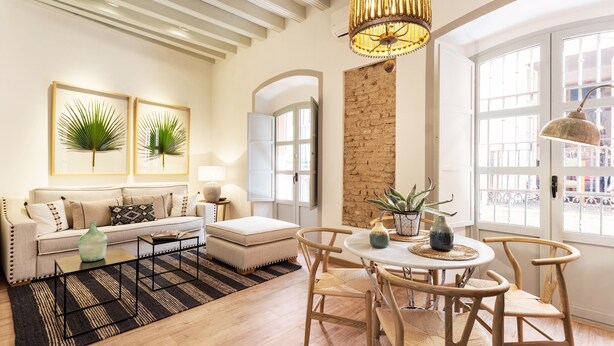 Una sala de estar moderna con sofá, reposapiés, mesa de centro y comedor con sillas. Se aprecia una araña, una planta y vistas al exterior a través de amplios ventanales.