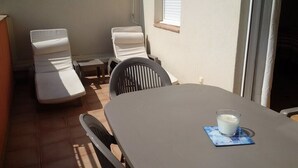 Un patio ensoleillé avec une table et deux fauteuils, un verre de lait posé sur la table, et une fenêtre avec des stores.