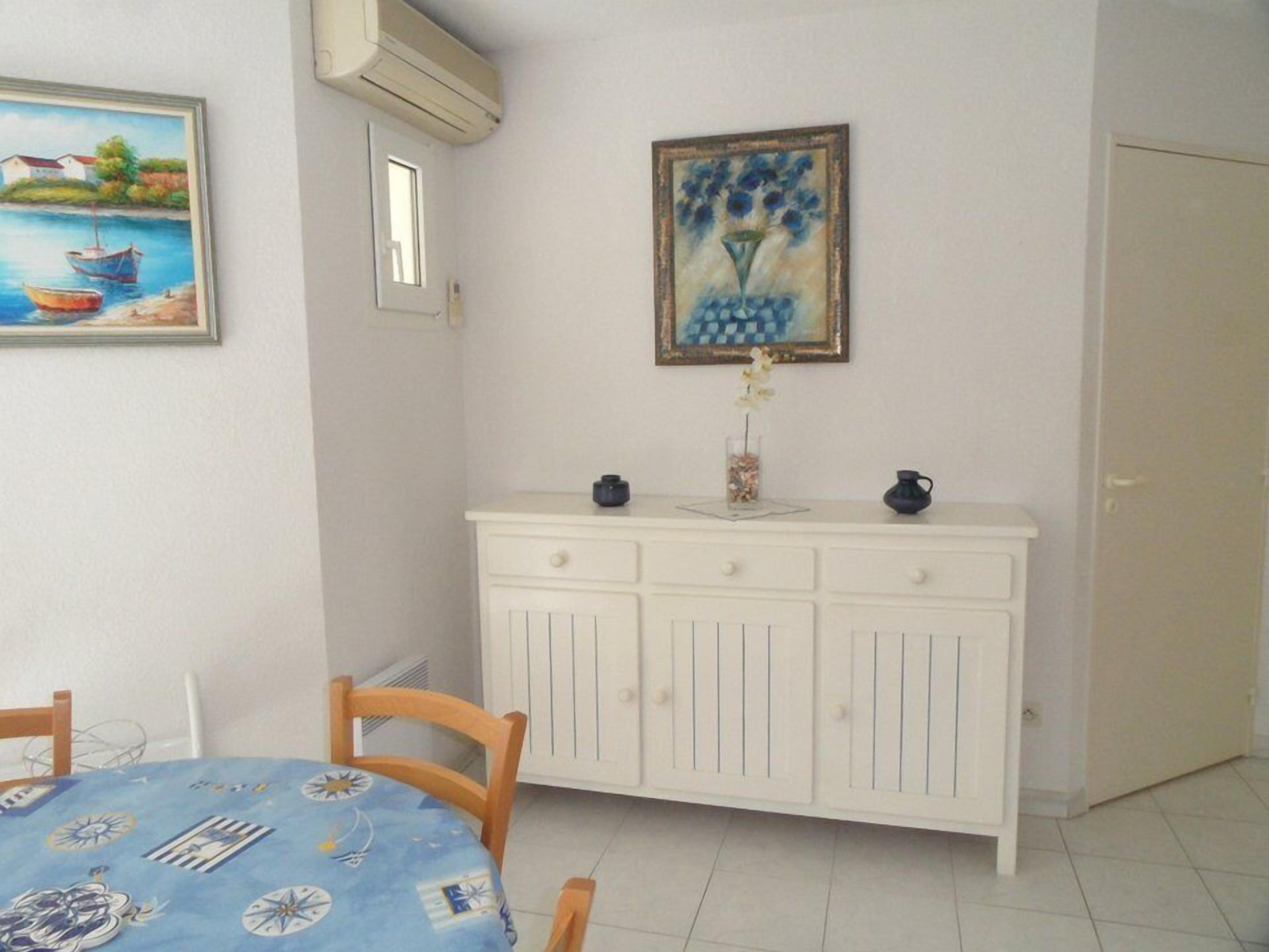Un área de comedor con una mesa preparada para dos personas, un armario blanco con dos cajones, y un cuadro de un jarrón azul con flores en la pared.