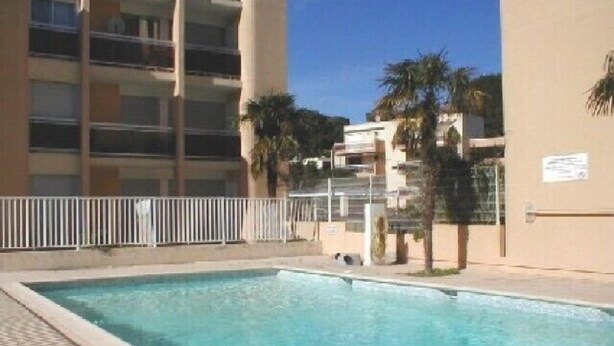 A swimming pool with a tiled deck, surrounded by a white fence, and adjacent to a multi-story building with balconies. - 2-room air-conditioned apartment, swimming pool (CAVALAIRE sur mer)