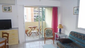 A living room with a sofa, a television, a dining table with chairs, a chair, a small table, and a sliding glass door leading to a balcony. - 2-room air-conditioned apartment, swimming pool (CAVALAIRE sur mer)