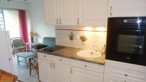 A kitchen with white cabinets, a black oven, and a sink. There is a chair and a table with a lamp in the background. - 2-room air-conditioned apartment, swimming pool (CAVALAIRE sur mer)