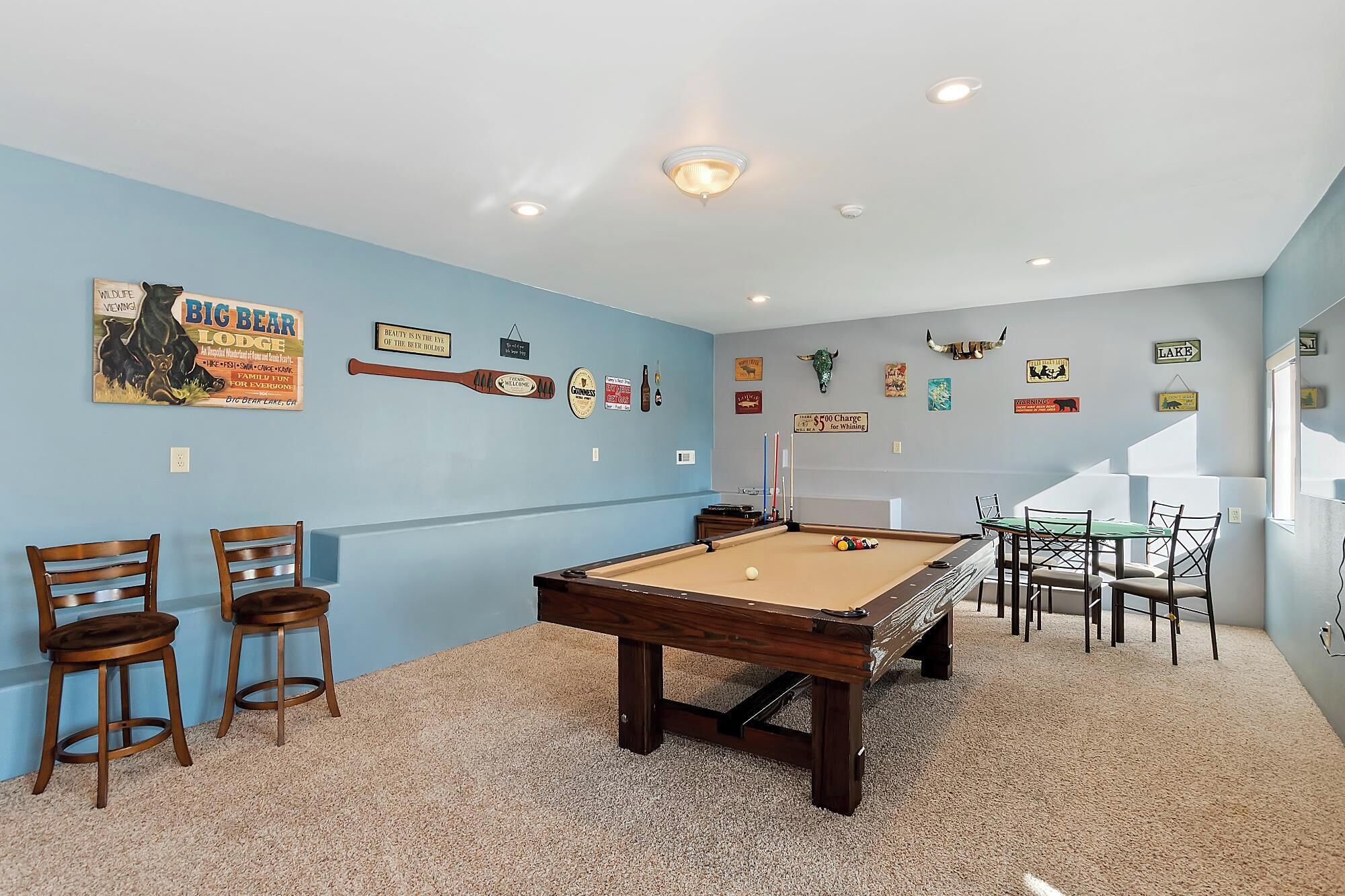 Una sala de billar con mesa de pool, sillas y varias decoraciones en las paredes, incluyendo un cartel con la leyenda 'Big Bear Lodge' y un cartel de 'Welcome'.