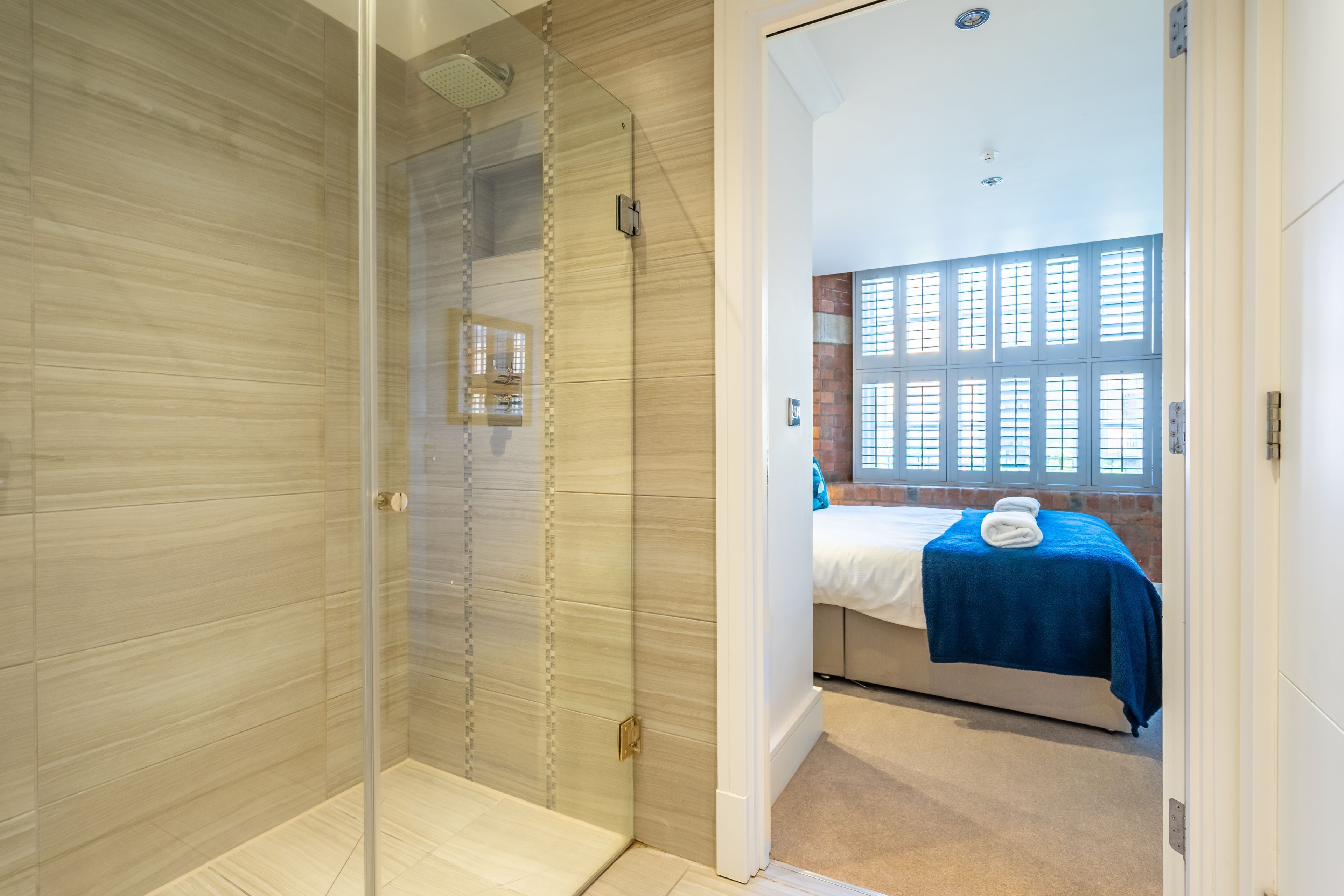 A modern bathroom with a glass shower enclosure, a visible towel rack, and a view of a bedroom with a bed and a window.
