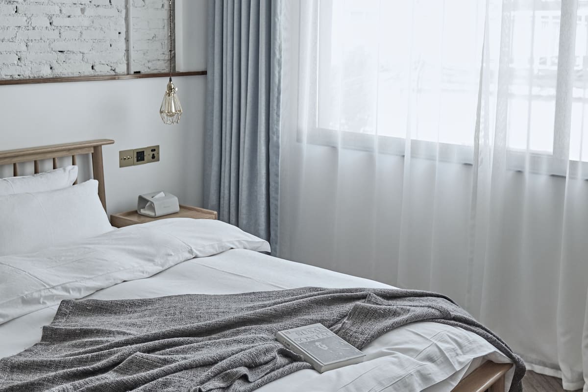 A bedroom with a wooden bed, a brick wall, a window with sheer curtains, and a hanging light fixture.