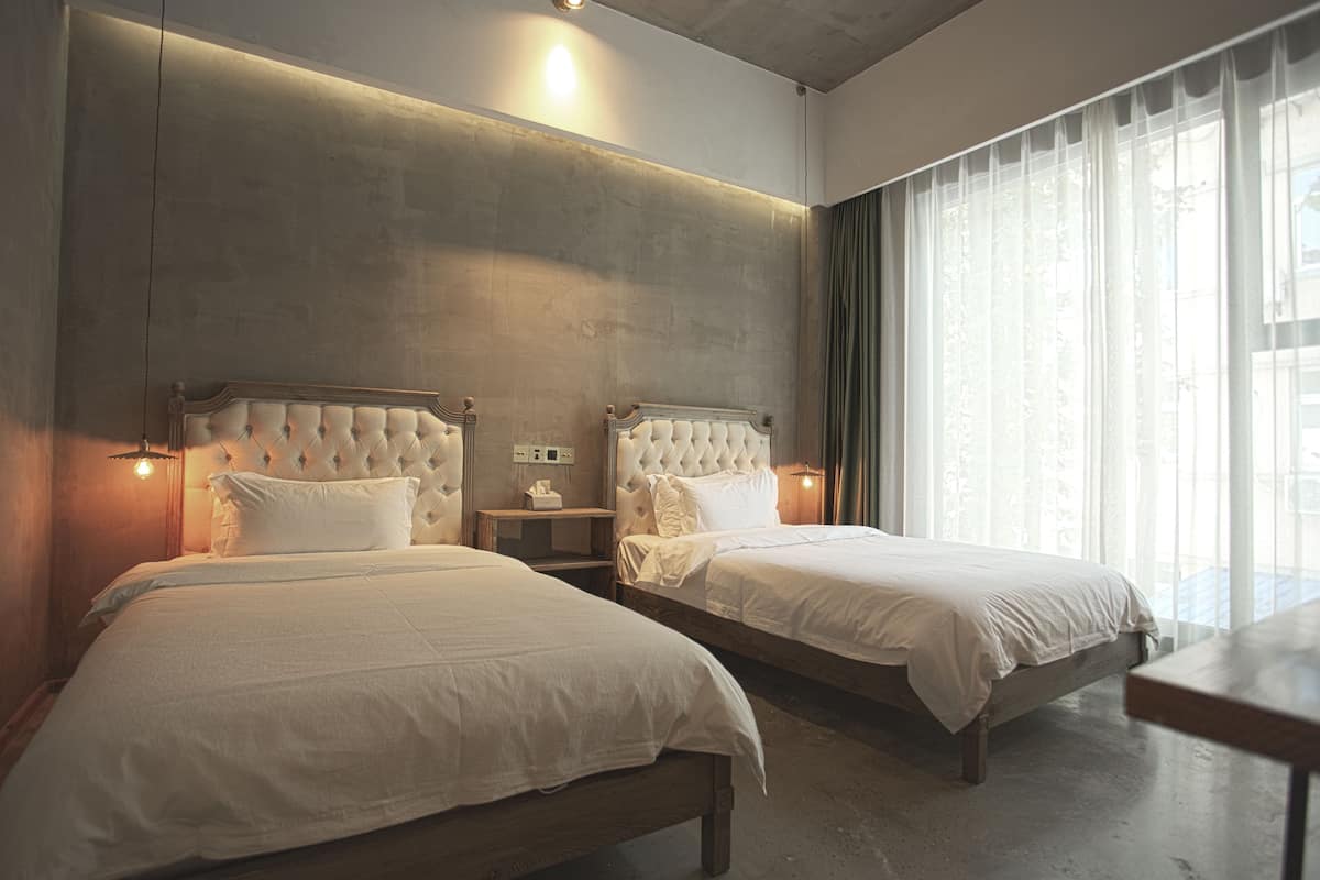 A hotel room with two beds, a wooden headboard, a bedside table with a lamp, and a large window with curtains.