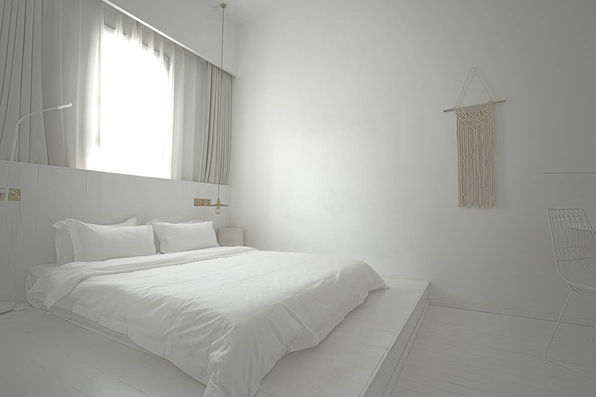 A minimalist white bedroom with a large bed, a bedside table, a hanging macrame wall decoration, and a white chair.