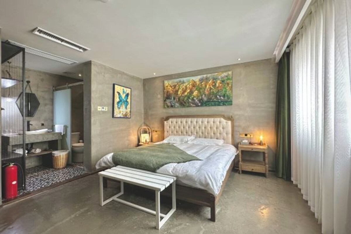 A bedroom with a bed, bedside tables, a painting, and a bathroom visible through an open door.