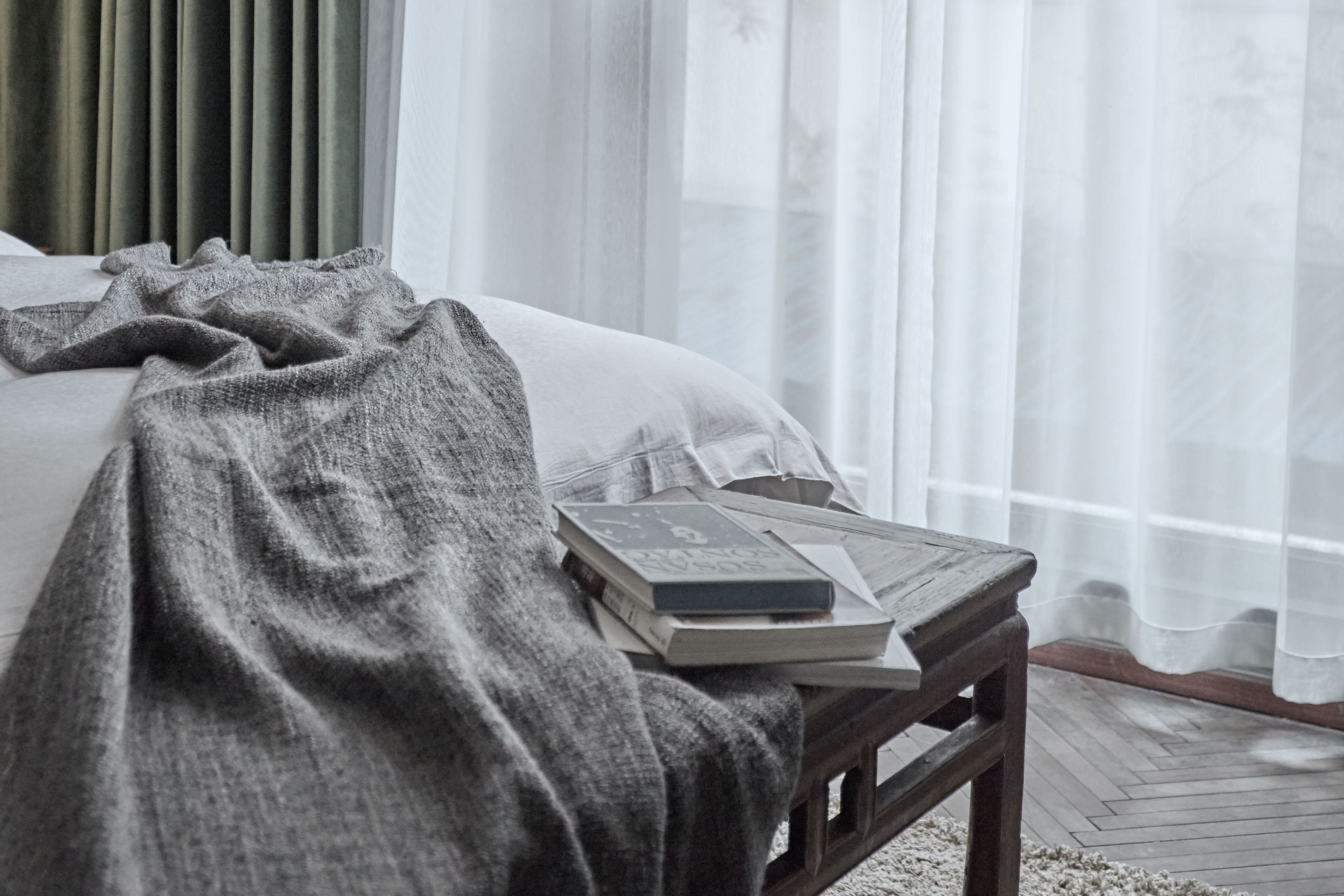 A neatly made bed with a grey blanket, a wooden bedside table with books, and sheer curtains.