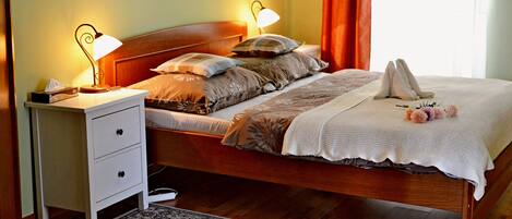 A neatly made bed with a white blanket and decorative pillows, a bedside table with a lamp, and a patterned rug on the wooden floor.