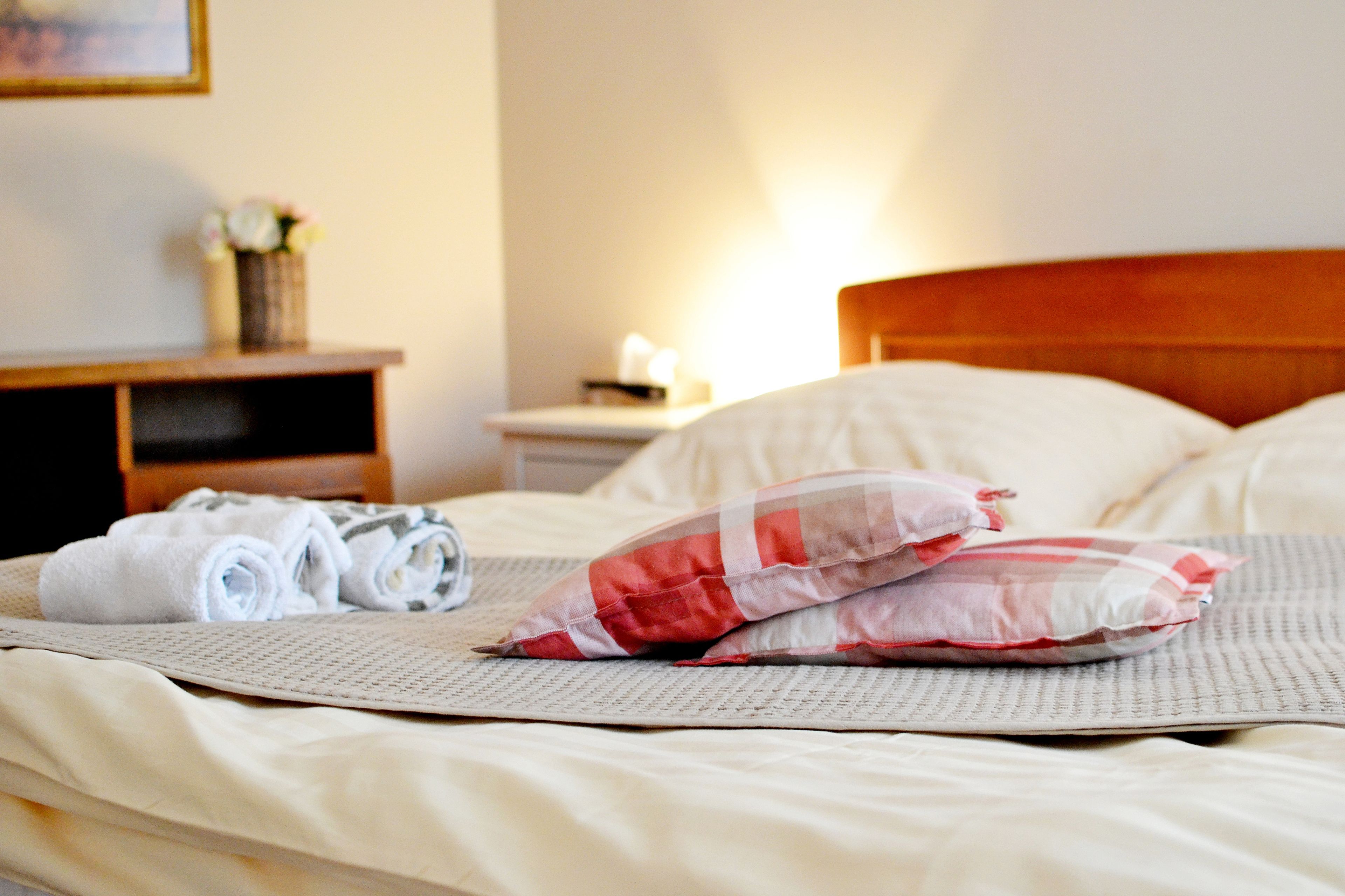 A neatly made bed with rolled towels and pillows, a bedside table with a vase of flowers, and a nightstand with a lamp.