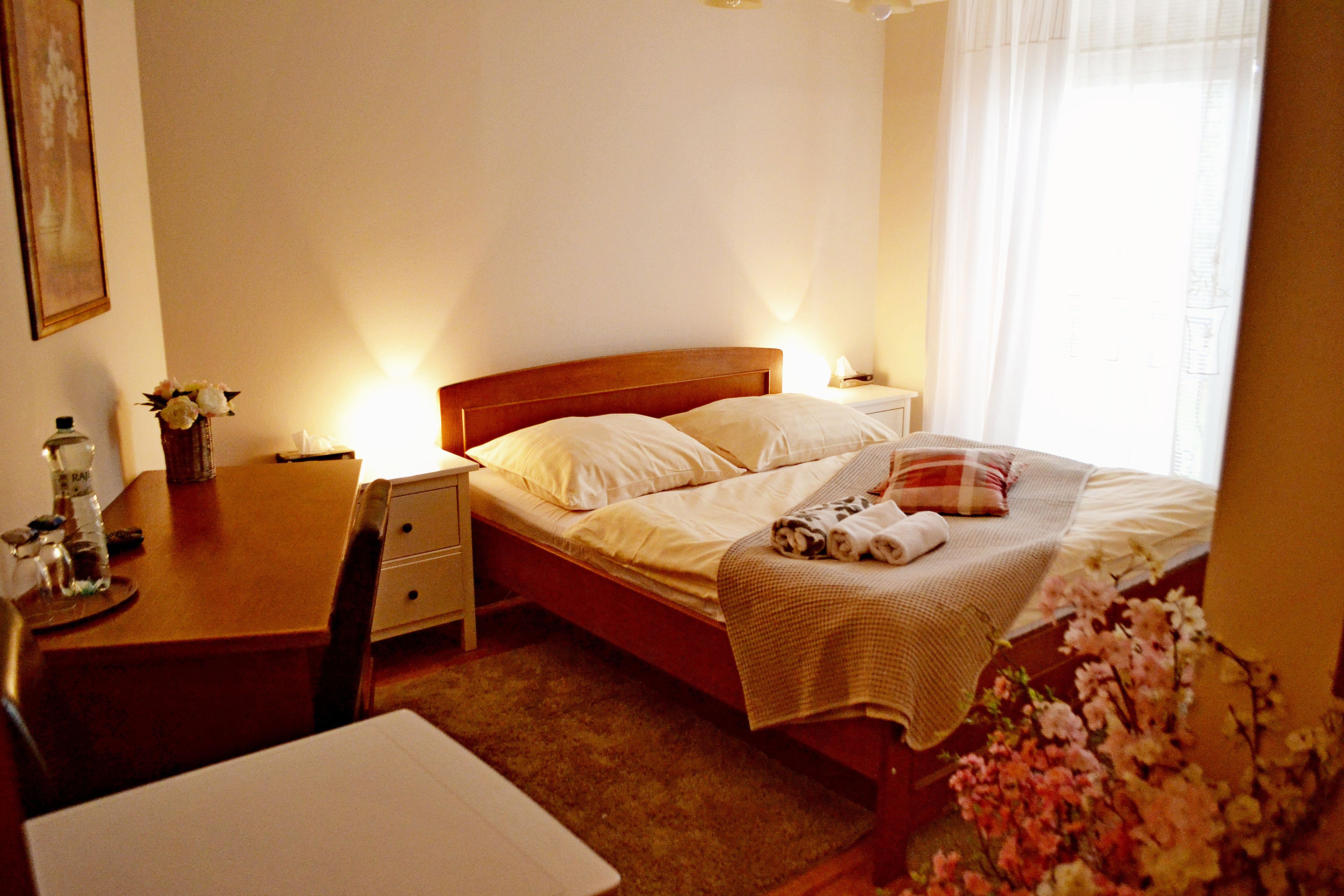 A neatly made bed with a blanket and pillows, a bedside table with a lamp, a chair, and a desk with a vase of flowers.