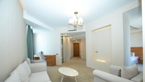 Un salon moderne avec un ensemble de canapés blancs, une petite table blanche et un lustre. On aperçoit un bureau en bois avec une chaise, et un rideau vert en arrière-plan.