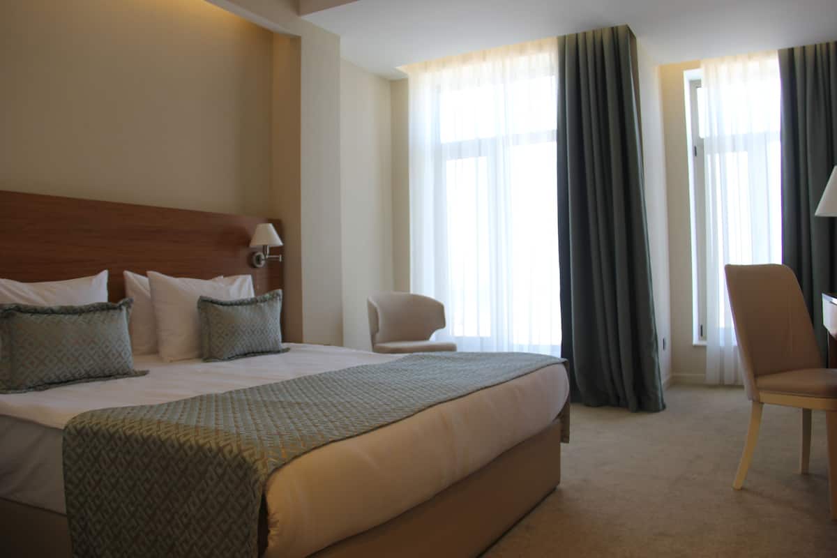 Une chambre d’hôtel avec un grand lit, deux chaises, un bureau et une fenêtre avec des rideaux.