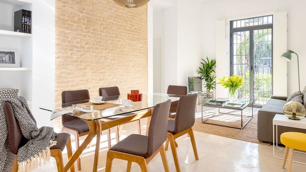 A modern living room with a glass dining table, comfortable seating, a brick accent wall, and a large window with a view. - Genteel Home Santa Teresa III (Sevilla)