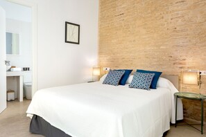 Une chambre avec un grand lit, une table de chevet avec une lampe, un mur en briques, une porte blanche et une petite salle de bain avec un lavabo et des toilettes.