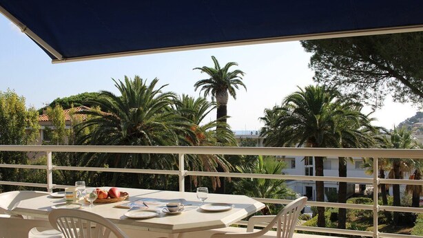 Ein Balkon mit Tisch für zwei Personen, mit Blick auf einen Garten mit Palmen und ein Gebäude im Hintergrund.