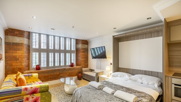A modern hotel room with a large window, a flat-screen TV, a bed with a grey duvet, a sofa with floral upholstery, and a small table. - Cocoa Suites 49 - York city centre studio (sleeps 2) (York)