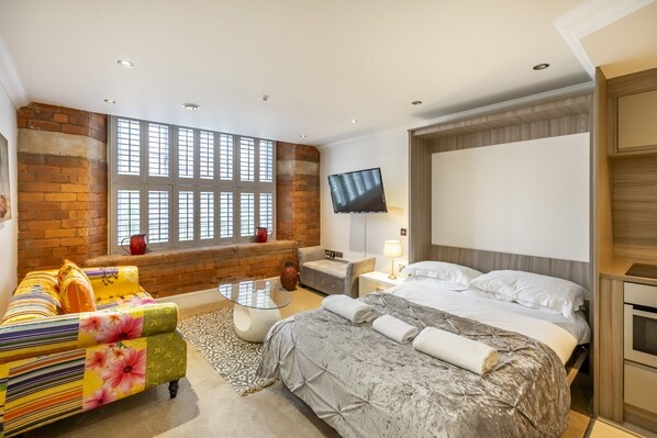 A modern hotel room with a large window, a flat-screen TV, a bed with a grey duvet, a sofa with floral upholstery, and a small table.