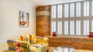 A room with a brick wall, a window with shutters, a sofa with floral upholstery, and a glass coffee table. - Cocoa Suites 49 - York city centre studio (sleeps 2) (York)