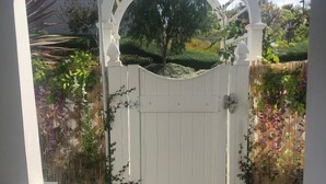 Um portão branco com um arco decorativo, dando acesso a um jardim com plantas e cerca.