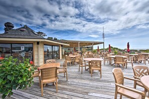 An outdoor patio area with wooden tables and chairs, a covered seating area, and a view of the sea. - Pool, Spa & Dining: Beach Condo in Hilton Head! (Hilton Head)
